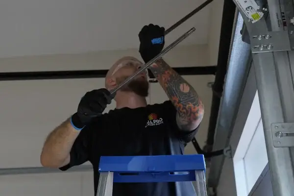 All Pro Garage Doors technician wearing a yellow hard hat and gloves carefully works with a tension rod on the torsion spring assembly above a white garage door.