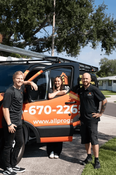 Three employees of All Pro Garage Doors—two men and one woman—standing proudly next to their bright orange company service van on a sunny suburban street.