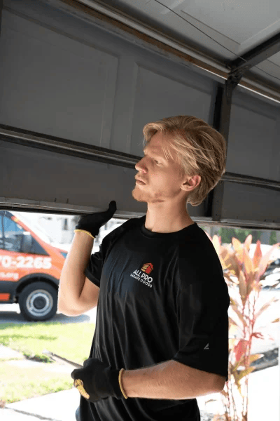 A close-up, vertical photograph of a young, blond All Pro Garage Doors technician wearing a black uniform shirt and gloves.